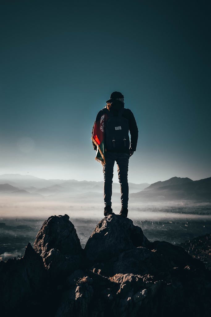 A lone hiker stands on a rocky peak, overlooking breathtaking mountains at sunrise in Afghanistan.